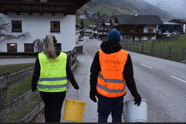 Zwei Menschen mit Warnwesten und Kübeln gehen die Straße runter