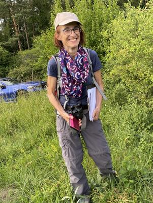 Frau mit Kappe und Unterlagen auf einer Wiese vor einer Hecke