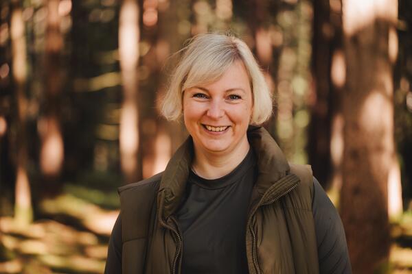 Portrait einer blonden Dame im Wald vor braunen Baumstämmen