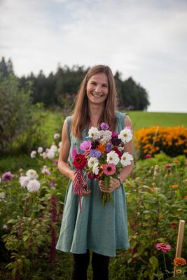 ine lächelnde Frau in einem hellblauen Kleid hält einen bunten Strauß selbstgepflückter Blumen – darunter Cosmos, Zinnien und Skabiosen – in einem blühenden Naturgarten mit Wald im Hintergrund.