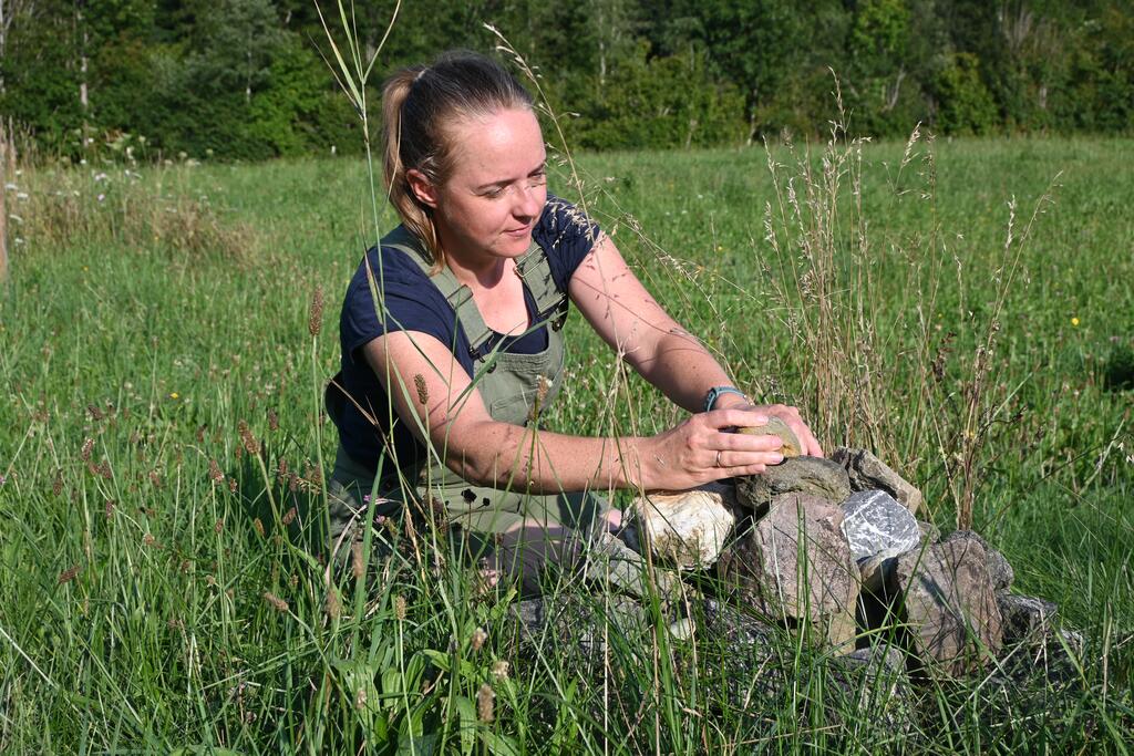 Eine Frau in grüner Latzhose kniet in einer Blumenwiese und legt eine Steinpyramide  an.