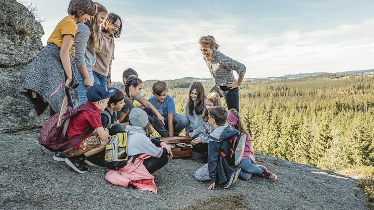 Eine Gruppe von Kindern, die sich auf einem Granitfelsen über den Baumwipfeln befindet, betrachtet eine Schatzkiste.