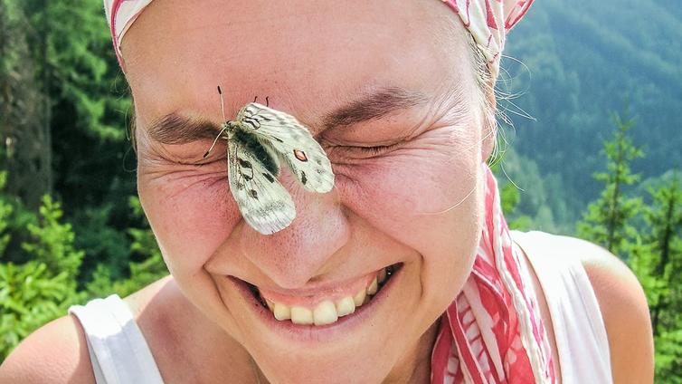 Ein Schmetterling sitzt im Gesicht einer jungen Frau, die darüber sehr amüsiert ist.