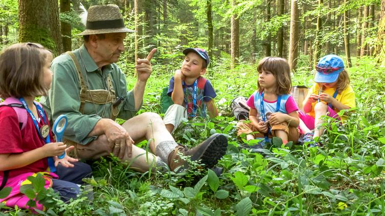 Der Schatz im Almtal © NATURSCHAUSPIEL, KinderUni OÖ Fritz Wolf erklärt vier Kindern auf dem Boden sitzend die Besonderheiten im Wald