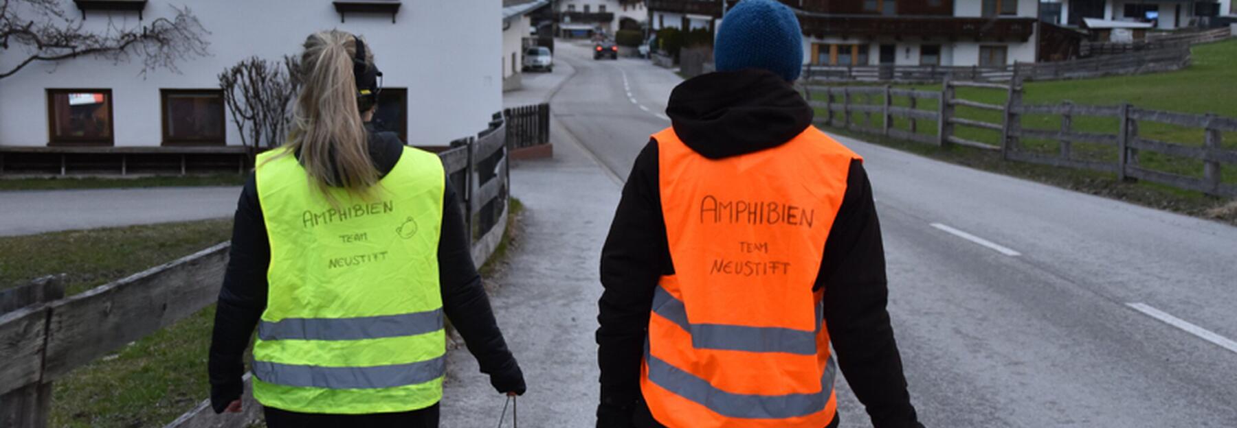 Zwei Menschen mit Warnwesten und Kübeln gehen die Straße runter