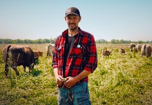Ein Mann steht vor vielen Kühen auf einer grünen Wiese 